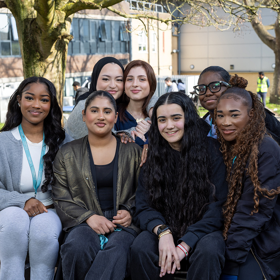 A group of female students sitting together in a group outside on a bench in the courtyard at Uxbridge College. They are smiling and there are trees and other students behind them. 