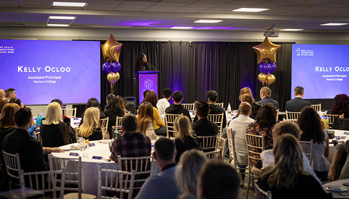 The audience at the HRUC Skills Competition award night listening to Kelly Ocloo, Assistant Principal at Harrow College. Kelly is standing on stage behind a lectern with large screens either side and balloon towers with gold stars on top. 