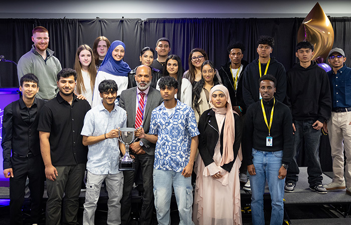 Uxbridge College students with their principal, Clive Hodge, holding the overall cup for the HRUC Skills Competition. They are on stage in a group and 2 male students at the front are holding a silver cup.