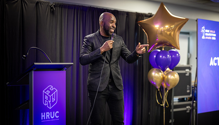 Action Jackson hosting the HRUC Skills Competition awards evening. He is standing on stage and pointing at the audience. 