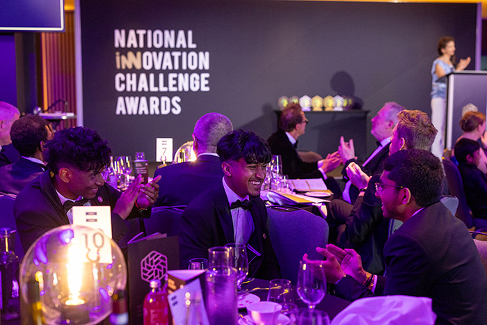 NIC 2026 colleges category winners - Shlok, Xainaldo and Faithon from Uxbridge College sitting at a table at the National Innovation Challenge awards ceremony. They are smiling and clapping and all dressed in formal wear.