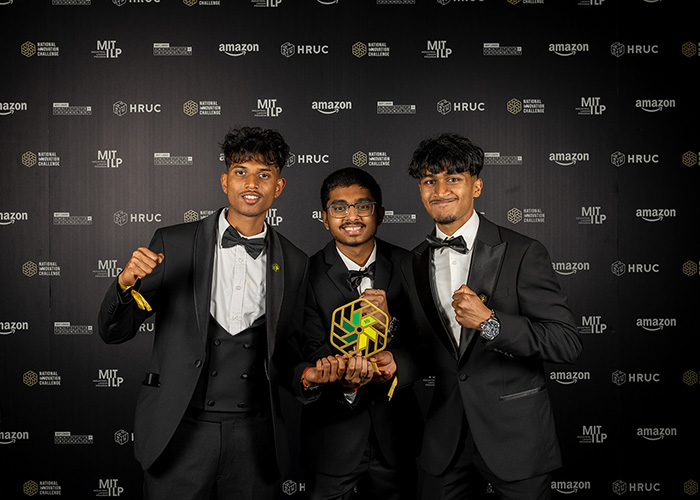 Shlok, Xainaldo and Faithon from Uxbridge College who were the winners of the National Innovation Challenge 2026. They are dressed in tuxedos, holding their award with fists held high. There is a black wall with white and gold logos behind them.