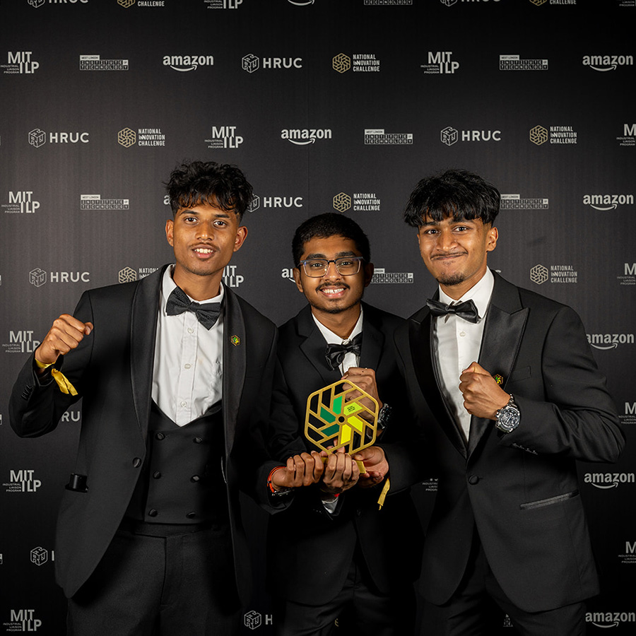 Shlok, Xainaldo and Faithon from Uxbridge College who were the winners of the National Innovation Challenge 2026. They are dressed in tuxedos, holding their award with fists held high. There is a black wall with white and gold logos behind them.