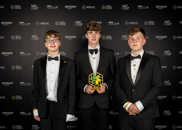 Timothy, Ollie and Wojtek from St Bede’s Catholic High School are the Secondary School Winners of the National Innovation Challenge 2026. They are dressed in tuxedos, holding their award and standing in front of a black wall with white and gold logos on it.