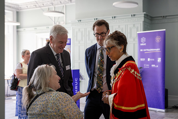 Mayor of Hillingdon Reeta Chamdal, talking with Keith Smith (CEO of HRUC) and others at the Barra Hall official opening event. Reeta is wearing her ceremonial robes and there is a Be Extraordinary HRUC purple banner behind them in the large room.  