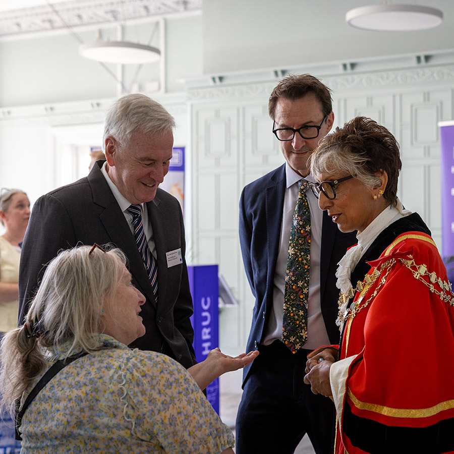 Mayor of Hillingdon Reeta Chamdal, talking with Keith Smith (CEO of HRUC) and others at the Barra Hall official opening event. Reeta is wearing her ceremonial robes and there is a Be Extraordinary HRUC purple banner behind them in the large room.  