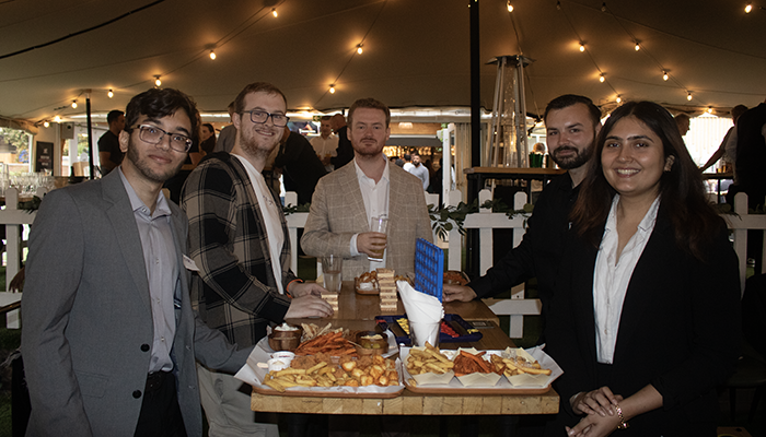 HRUC Alumni at the Finsbury Square Garden Bar at the first alumni networking event. They are standing around a table and there is food drinks in front of them. They are under an outdoor tent area. 