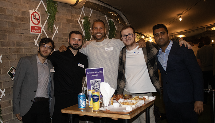 Ryan Rankin, Uxbridge College alumnus, in a group with other male alumnus. Their arms are around each other's shoulders. There is a brick wall behind them and food and drinks on a table in front of them.