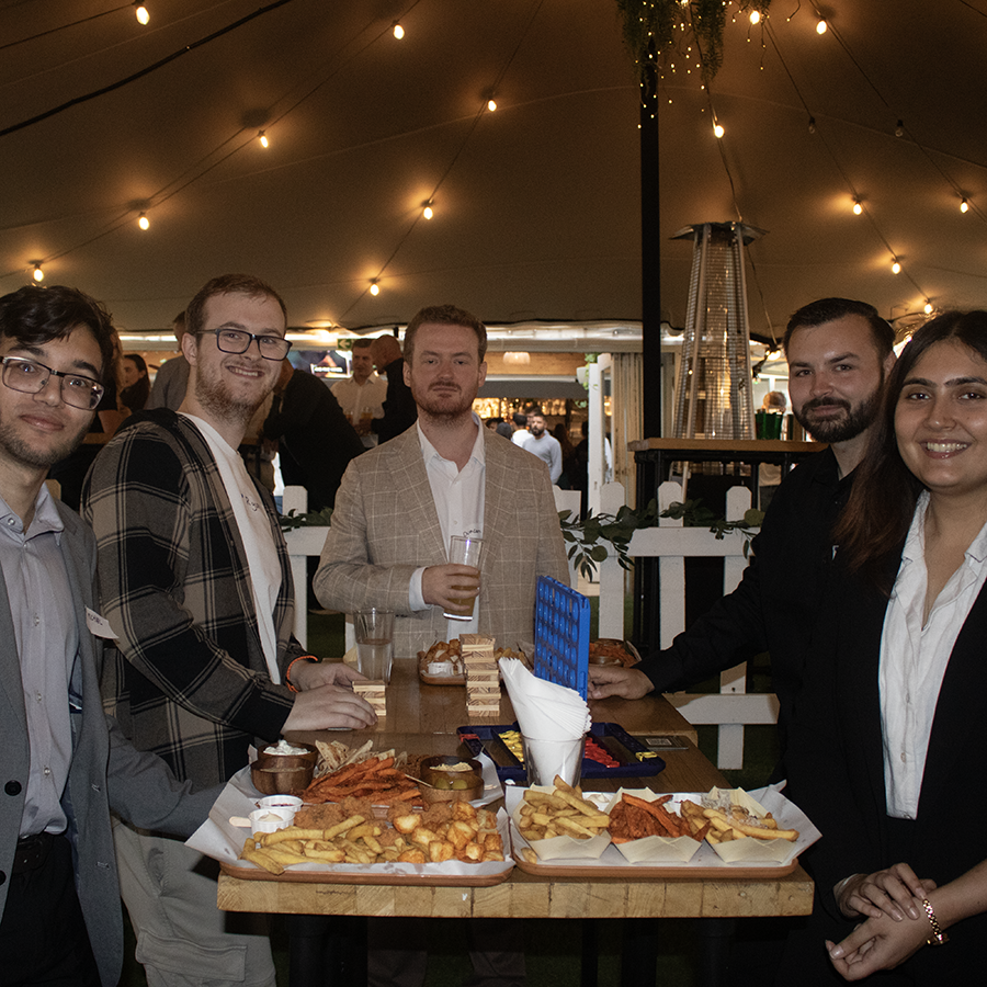 HRUC Alumni at the Finsbury Square Garden Bar at the first alumni networking event. They are standing around a table and there is food drinks in front of them. They are under an outdoor tent area. 