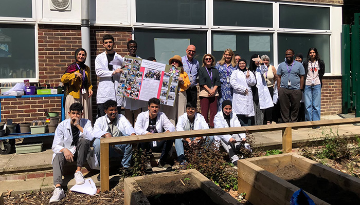 Harrow College ESOL students with teaching staff gathered in front of the Northwood Hills Underground community garden project. Some are wearing white lab coats to over their clothes. 