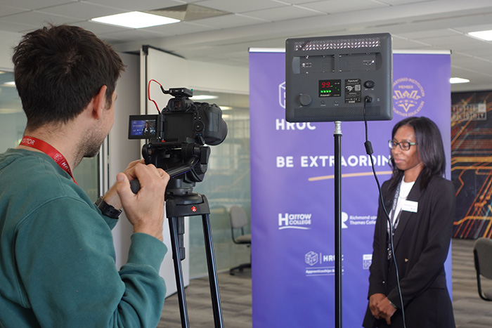 Camera man shooting video of a student interview. The female student is standing in front of a purple Be Extraordinary branded banner and there is a light in the foreground pointed towards her.       
