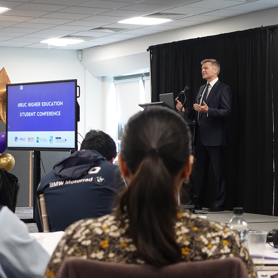 Scott Gregory; success coach and author; giving a keynote session at the Higher Education Student Conference. There are students sitting at tables in front of him; with screens to his left and he is standing behind a lectern.       
