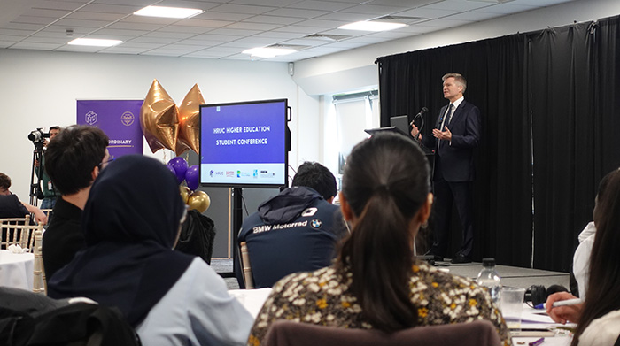 Scott Gregory; success coach and author; giving a keynote session at the Higher Education Student Conference. There are students sitting at tables in front of him; with screens to his left and he is standing behind a lectern.       