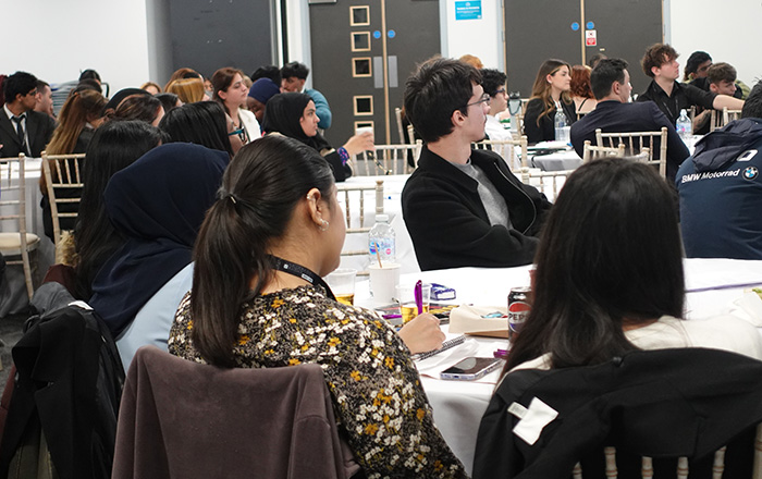 Students sitting at tables at the Higher Education Student Conference. They are listening to a speaker on stage.     