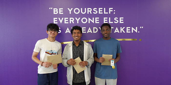 Three male Uxbridge College students holding their GCSE exam results envelopes and standing in front of a purple wall with the quote from Oscar Wilde that says: Be yourself; everyone else is already taken.        