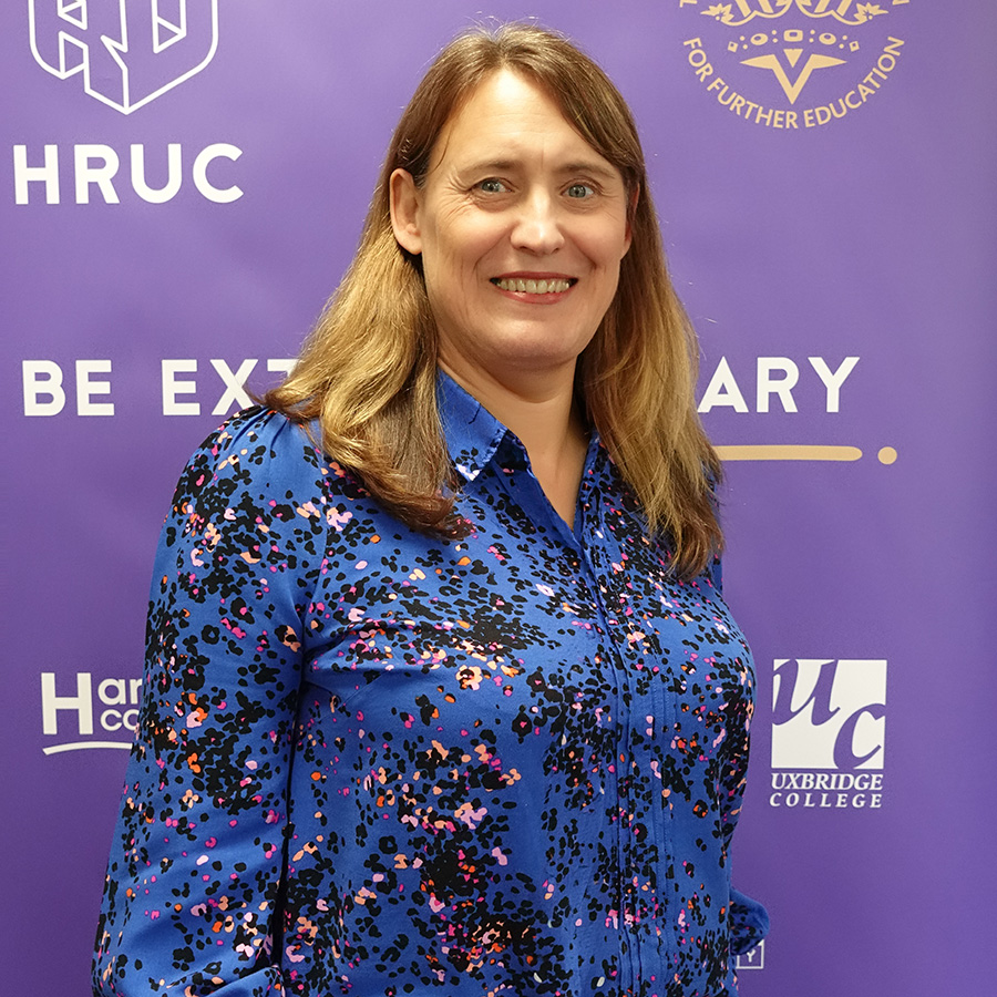 Jo Swindells, new Principal of Harrow College - she is wearing a blue top with black, white and pink spots, has shoulder length brown hair and is standing in front of an HRUC Be Extraordinary banner.