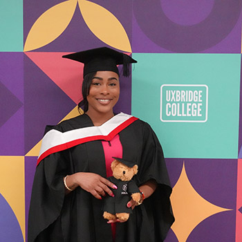 Female HE graduate in her black cap and gown dressed for her gradution ceremony, with a red and white sash over her shoulders and holding a teddy bear dressed in black cap and gown. She is standing in front of the Uxbridge College new logo and branding colours. 