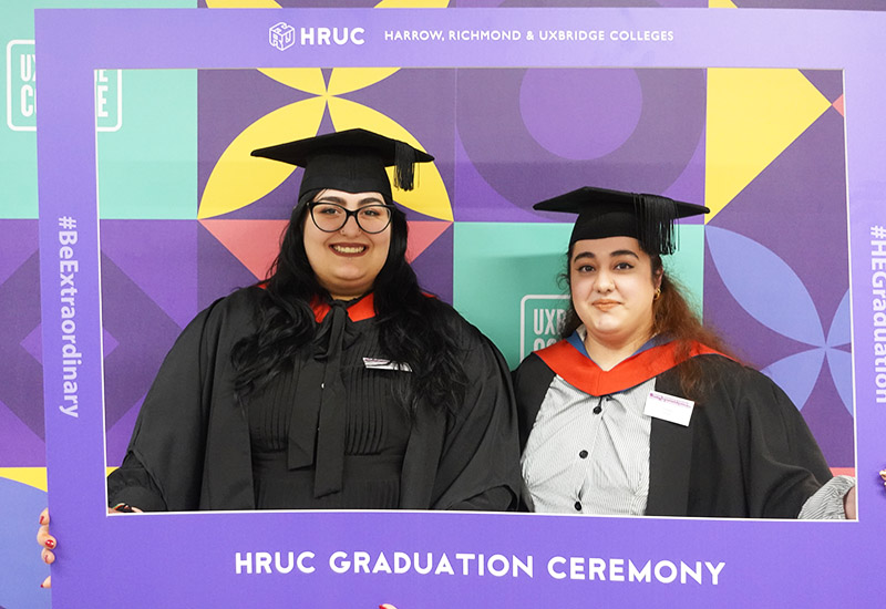 Two female HE graduates in their black caps and gowns dressed for their gradution ceremony. Both with a red sash over their shoulders.