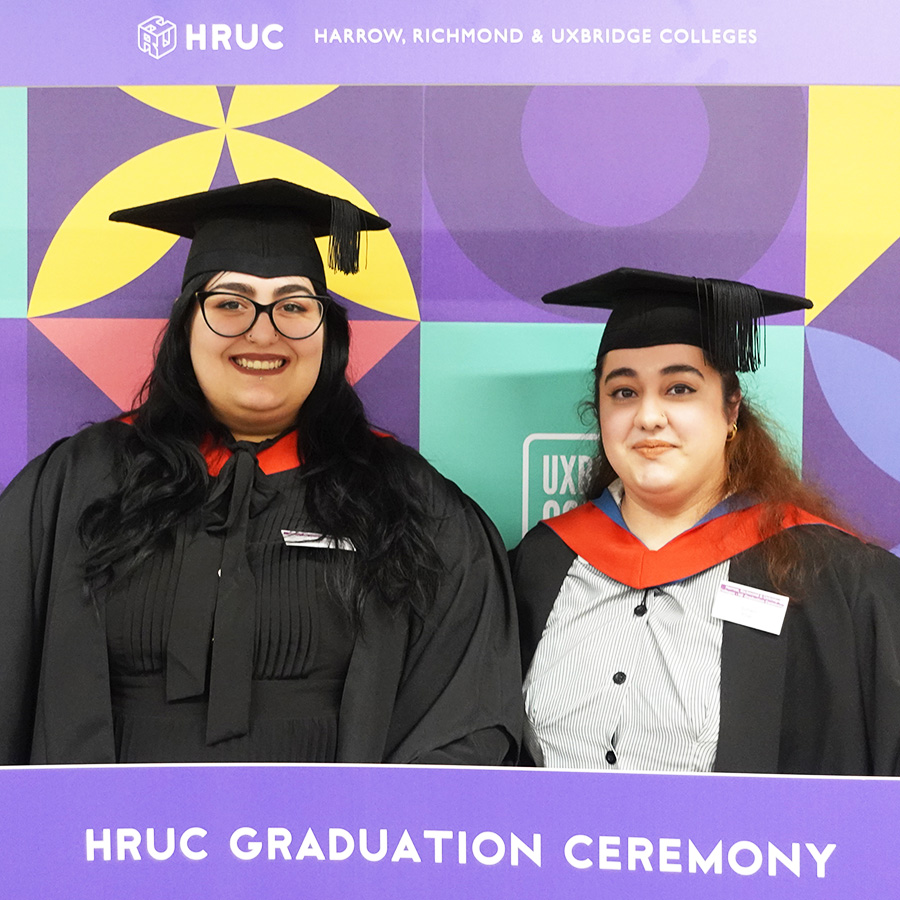 Two female HE graduates in their black caps and gowns dressed for their gradution ceremony. Both with a red sash over their shoulders.