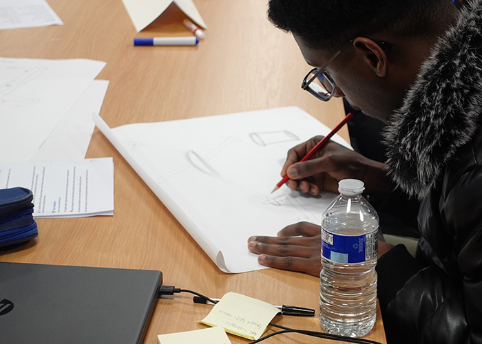 A student is working on designing a concept as part of the Real-World Amazon Challenge in Innovative Mini Project. He is drawing on paper and there is a laptop and water bottle in the foreground.