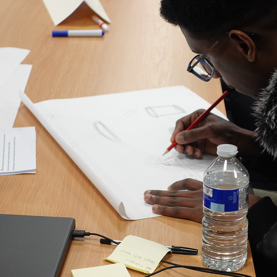 A student is working on designing a concept as part of the Real-World Amazon Challenge in Innovative Mini Project. He is drawing on paper and there is a laptop and water bottle in the foreground.