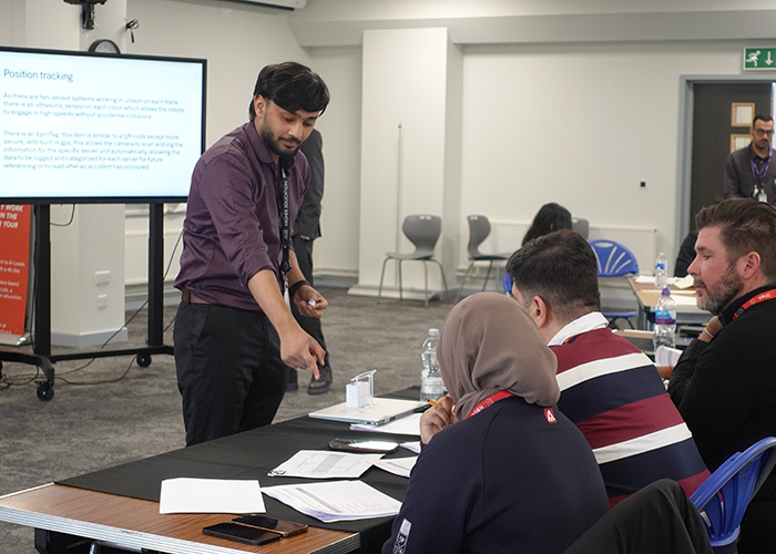 A staff member is helping out a team at the Real-World Amazon Challenge in Innovative Mini Project - they are pointing to a paper and three of the team are looking at it. They are in a classroom and there is a screen behind the staff member with a slide on it that is titled: Position tracking.    