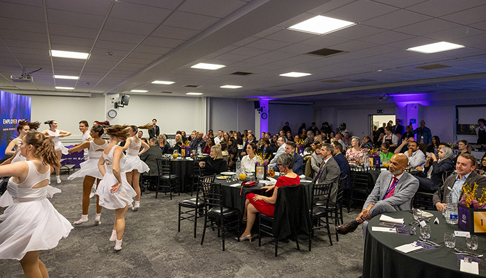 Praxis performers dancing in front of the audience at the Employers Awards evening. The performers are all wearing white and the audience are sitting around tables with black table cloths.