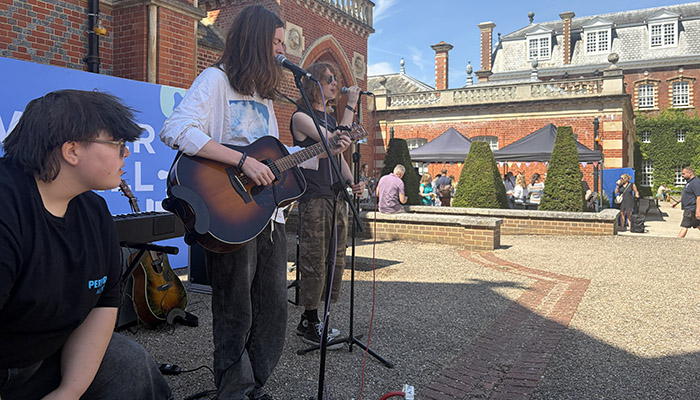 Uxbridge College students performing at the Festival of Education. They are standing behind microphones and the lead singer is playing guitar, while another is playing a percussion box. Wellington College grounds and buildings are in the background.