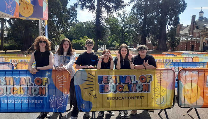 Uxbridge College students standing at the barriers of the Festival of Education held at Wellington College. There are tall pine trees behind them and buildings. They are almost all wearing black tops with one in a long sleeve white top.