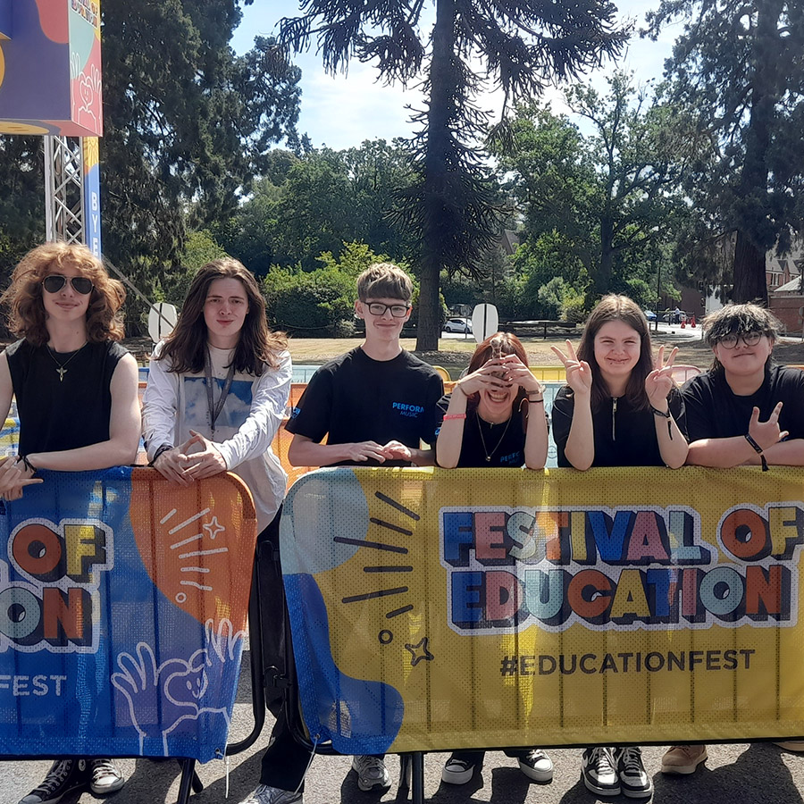 Uxbridge College students standing at the barriers of the Festival of Education held at Wellington College. There are tall pine trees behind them and buildings. They are almost all wearing black tops with one in a long sleeve white top.