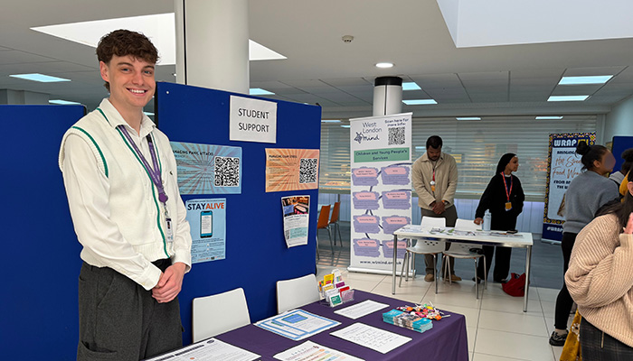 An HRUC staff member at the Mental Health Fair. He is smiling and standing behind a table with pamphlets and information and there are blue boards behind him with student support information.