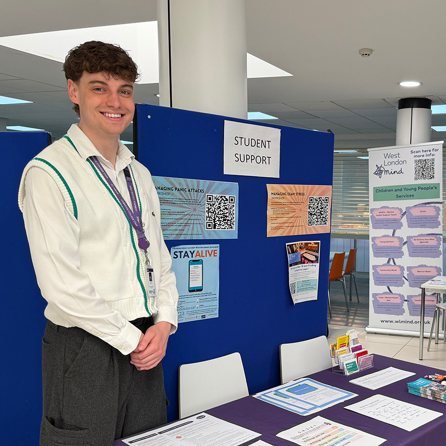 An HRUC staff member at the Mental Health Fair. He is smiling and standing behind a table with pamphlets and information and there are blue boards behind him with student support information.