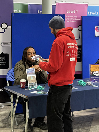 A student talking to a staff member at the HRUC Mental Health Fair. The student is wearing a bright red hoodie and holding leaflets of information.  