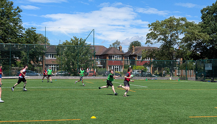 HRUC students playing rugby on an outside field. They are wearing red and green bibs. Sky is very blue with clouds and the grass is very green.