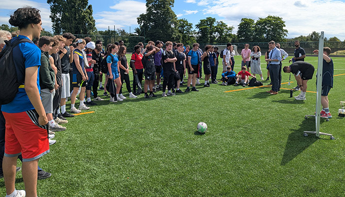 Students gathered on a sports field on HRUC Sports Day listenig to Gavin Hughes, Principal for Richmond upon Thames College. There is a football in the middle in front of them. They are all dressed in sports gear and there is a board on wheels in front of them.