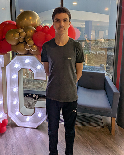 Male Harrow College student standing in Harrow on the Hill College reception after receving his GCSE exam results. There is a lit up sign behind him with the letter C visible and red and gold balloons on top of the sign. He is wearing a grey t-shirt and blue track suit pants.