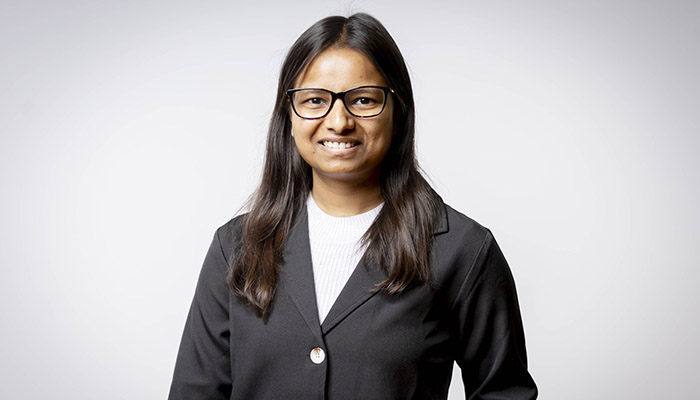 Pragna Navinkumar - former Harrow College student. She is wearing a black jacket and white top and has glasses with long brown hair. 