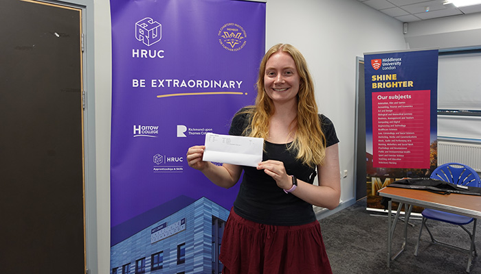 Uxbridge College student Rebecca holding her A-Level results in her hand and standing in front of a Be Extraordinary banner. She looks very happy.     
