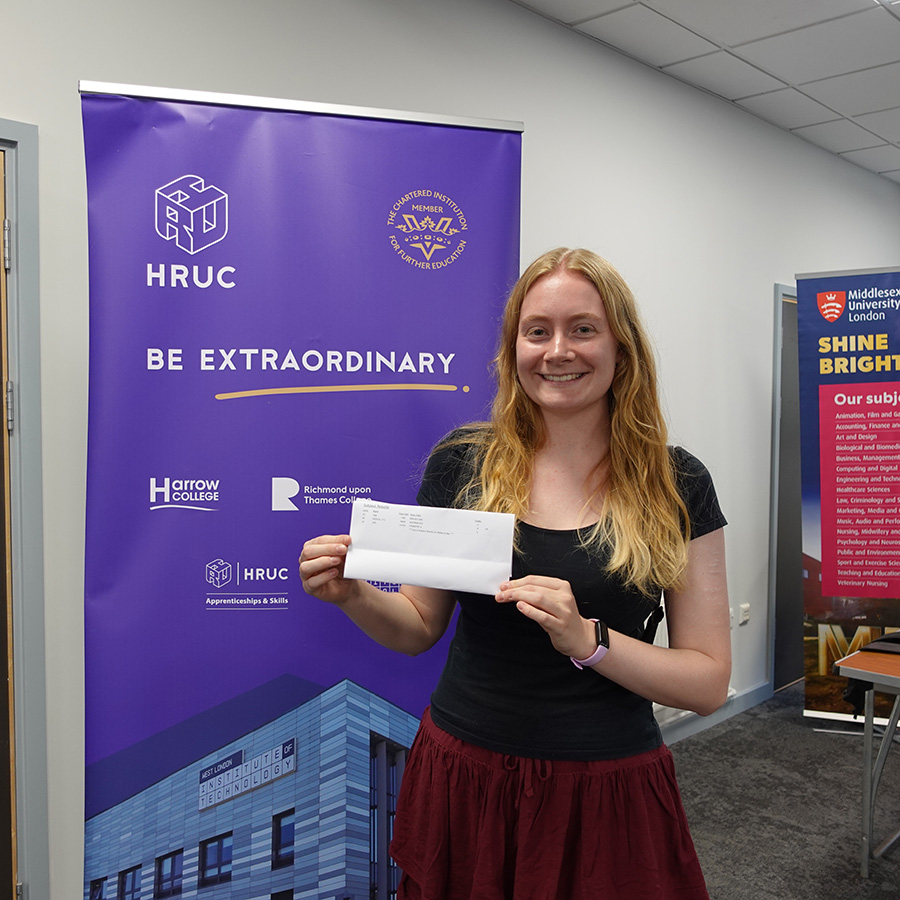 Uxbridge College student Rebecca holding her A-Level results in her hand and standing in front of a Be Extraordinary banner. She looks very happy.     