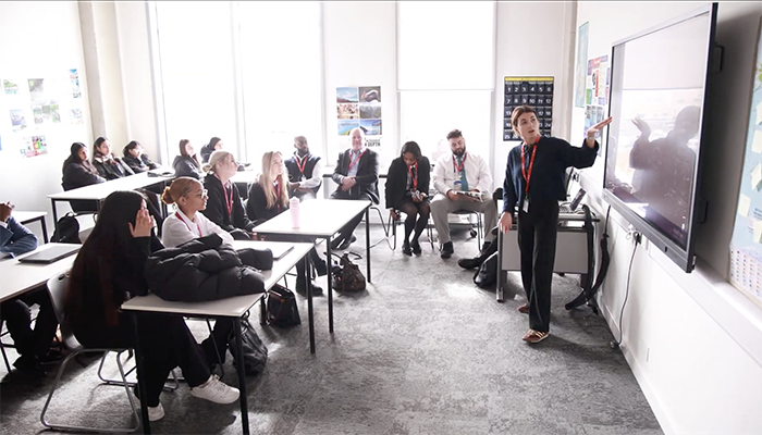 HRUC students being taught the Skills Builder Universal Framework. They are sitting in a classroom and listening to a teacher at the front of the class who is pointing to a screen. There are windows with bright sunlight streaming in to the left of them. 
