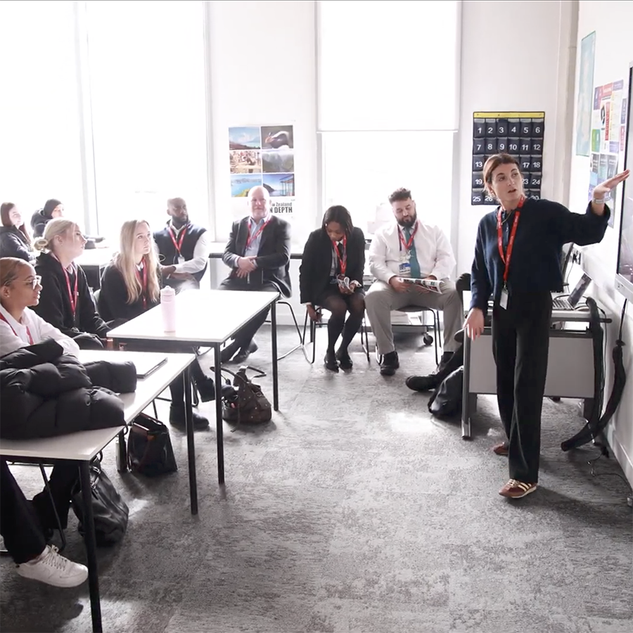 HRUC students being taught the Skills Builder Universal Framework. They are sitting in a classroom and listening to a teacher at the front of the class who is pointing to a screen. There are windows with bright sunlight streaming in to the left of them. 