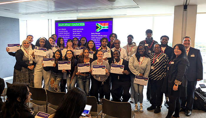 HRUC's Turing Scheme students who went to South Africa - they are standing in a group, smiling and holding their certificates with HRUC staff members either side of them. Behind them is a screen with a slide with all their names on it and floor to ceiling windows. 