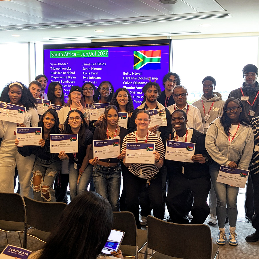 HRUC's Turing Scheme students who went to South Africa - they are standing in a group, smiling and holding their certificates with HRUC staff members either side of them. Behind them is a screen with a slide with all their names on it and floor to ceiling windows. 