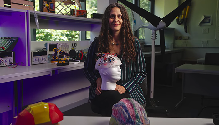 Valentina Vicario sitting in an engineering classroom with her cranial cooler prototype on her lap. She is wearing a striped jacket and has long brown curly hair.