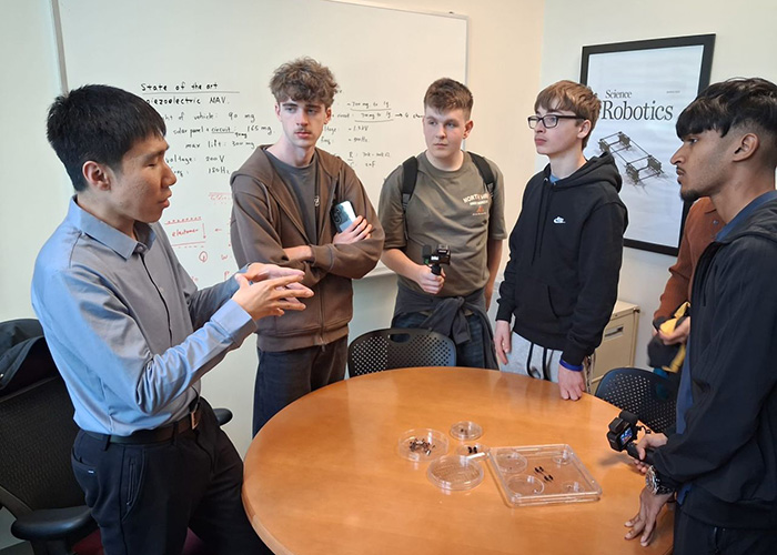 Our NIC 2026 winners listening to an MIT staff member. They are standing around a table and there is a whiteboard behind them with the words: State of the Art on it and a poster about robotics.