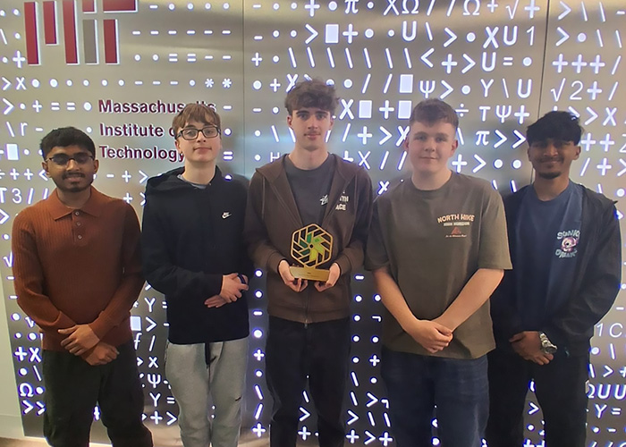 Our NIC 2026 winners standing in front of a Massachusetts Institute of Technology (MIT) metal sign and wall with maths symbols all over it. The middle boy is holding the NIC gold trophy.