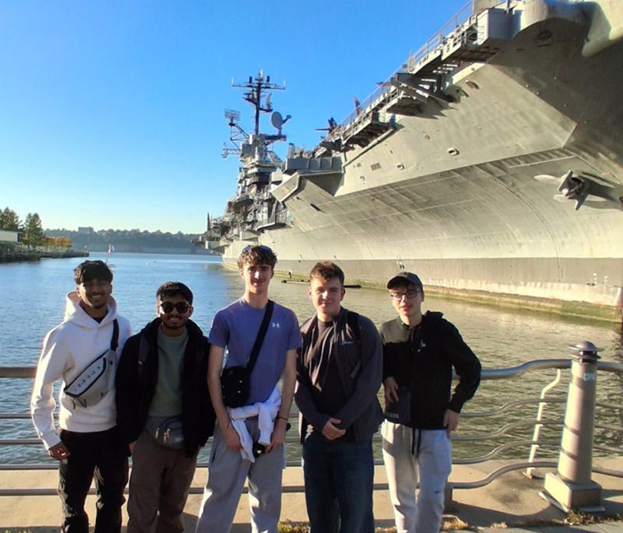 Our NIC 2026 winners standing in front of an aircraft carrier that is docked. The railing is behind them and it is a sunny day with bright blue sky.