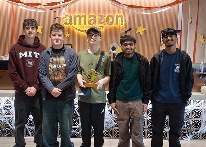 Our NIC 2026 winners standing in front of Amazon reception area with the Amazon sign behind them in yellow and Halloween decorations are displayed. They are holding the NIC gold trophy.