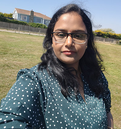 Kavitha - Electrical/Electronic Engineering Technician at Richmond upon Thames College. She is outside with green grass behind her, wearing a green top with white spots and glasses.