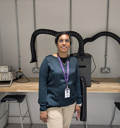 Pradeepa Sundaram, Engineering Lecturer at Richmond upon Thames College - she is stnading in front of a bench with equipment on it and large black tubes coming out of it. She is wearing glasses with a teal long sleeve top and cream trousers and a purple lanyard around her neck.